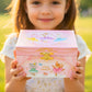 Child holding a pink jewelry box with ballerina designs outdoors.