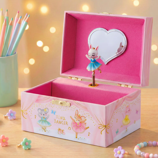 Pink music box with dancing ballerina design on a wooden surface with colorful pencils and flowers.