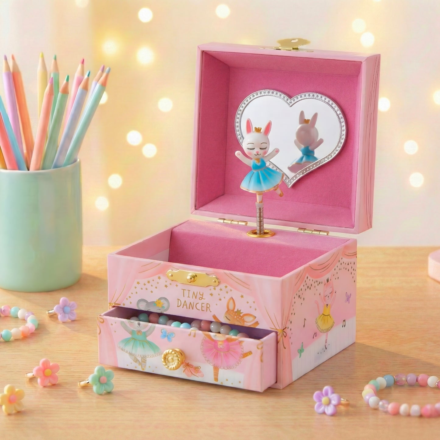 Pink music box with dancer design on a wooden surface with colorful pencils and flowers.