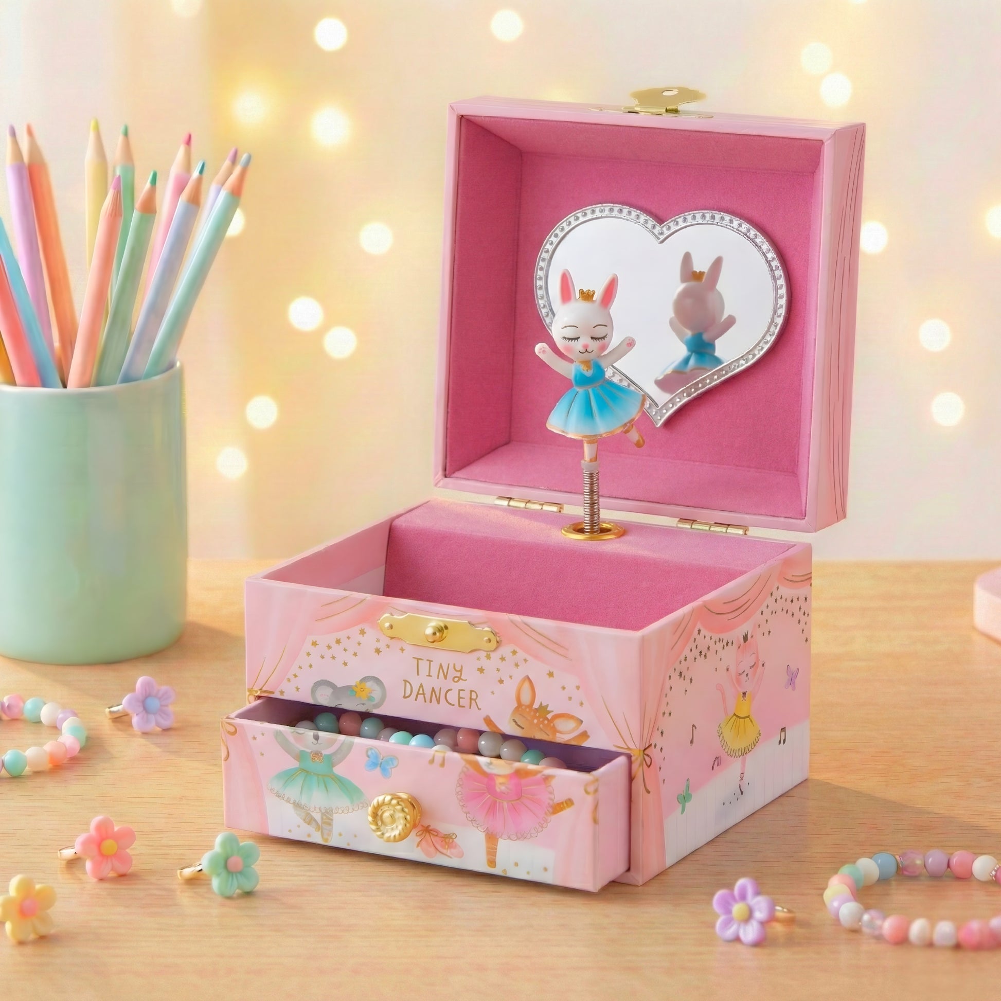 Pink music box with dancer design on a wooden surface with colorful pencils and flowers.