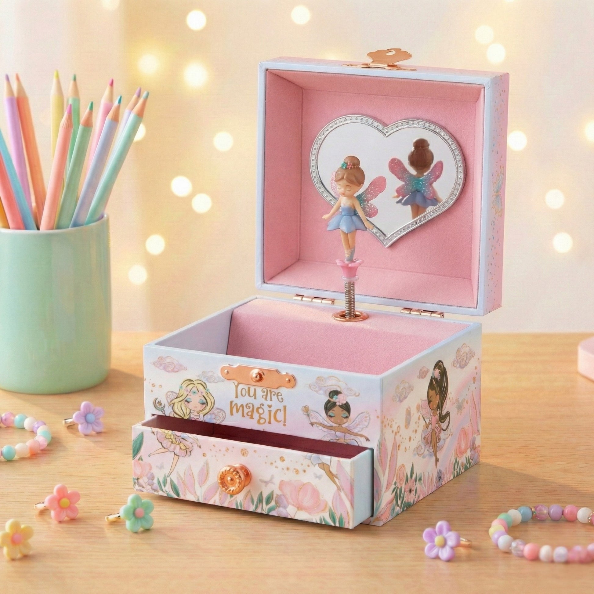 Pink fairy-themed music box with a drawer on a wooden surface with colorful pencils and flowers.