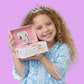 Young girl in a blue dress with a tiara holding a pink jewelry box against a purple background