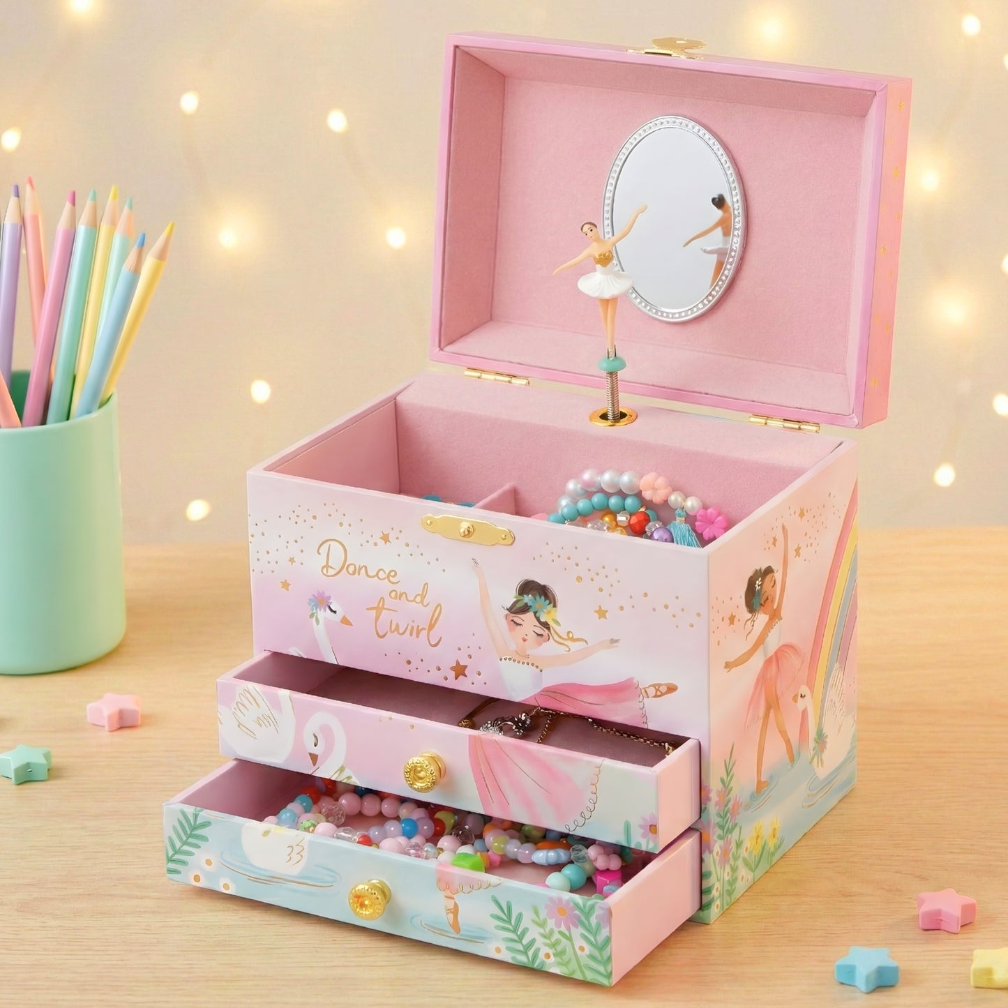 Pink jewelry box with ballerina design on a wooden surface with colorful pencils and beads.