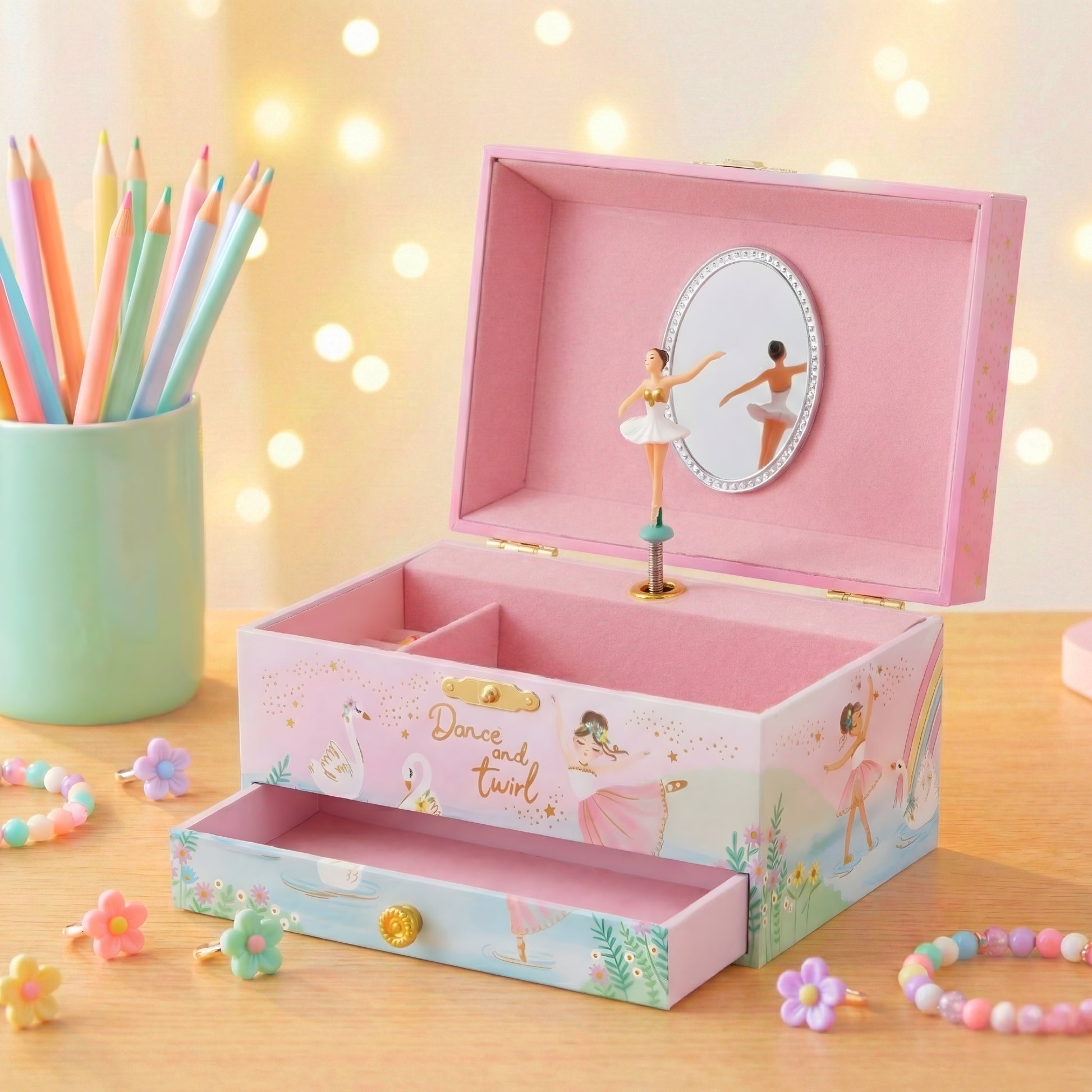 Pink music box with ballerina design on a wooden surface with colorful pencils and flowers.