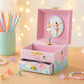 Pink music box with ballerina design on a wooden surface with colorful pencils and bracelets.