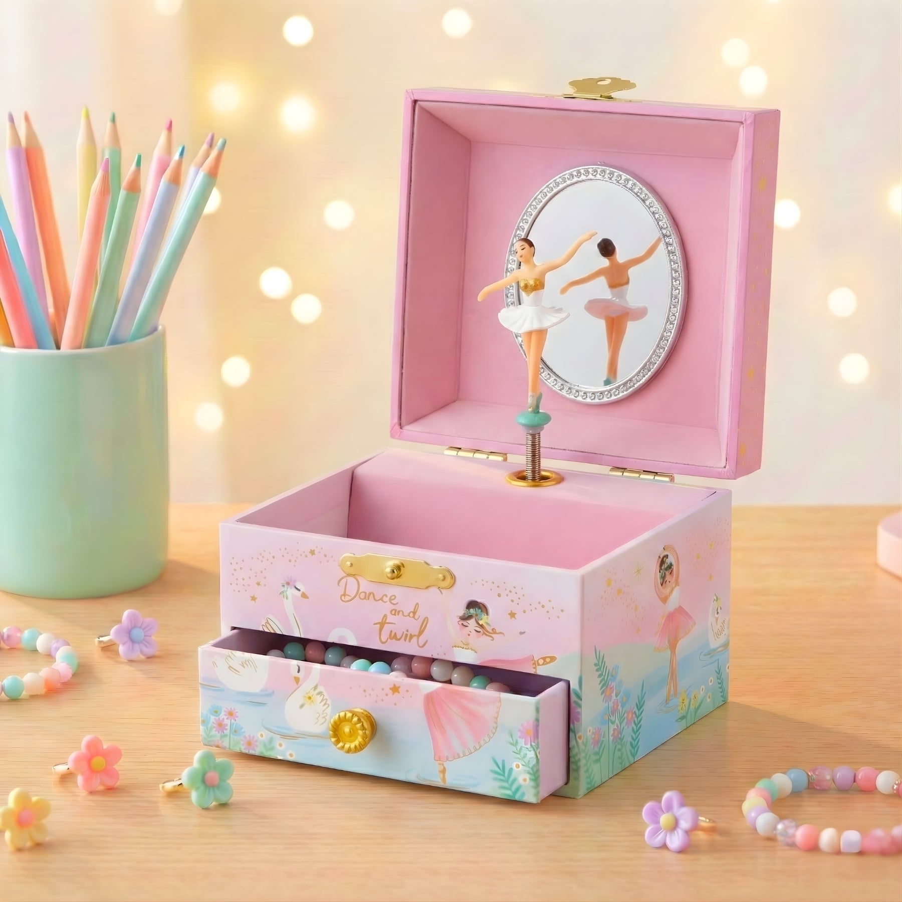 Pink music box with ballerina design on a wooden surface with colorful pencils and bracelets.