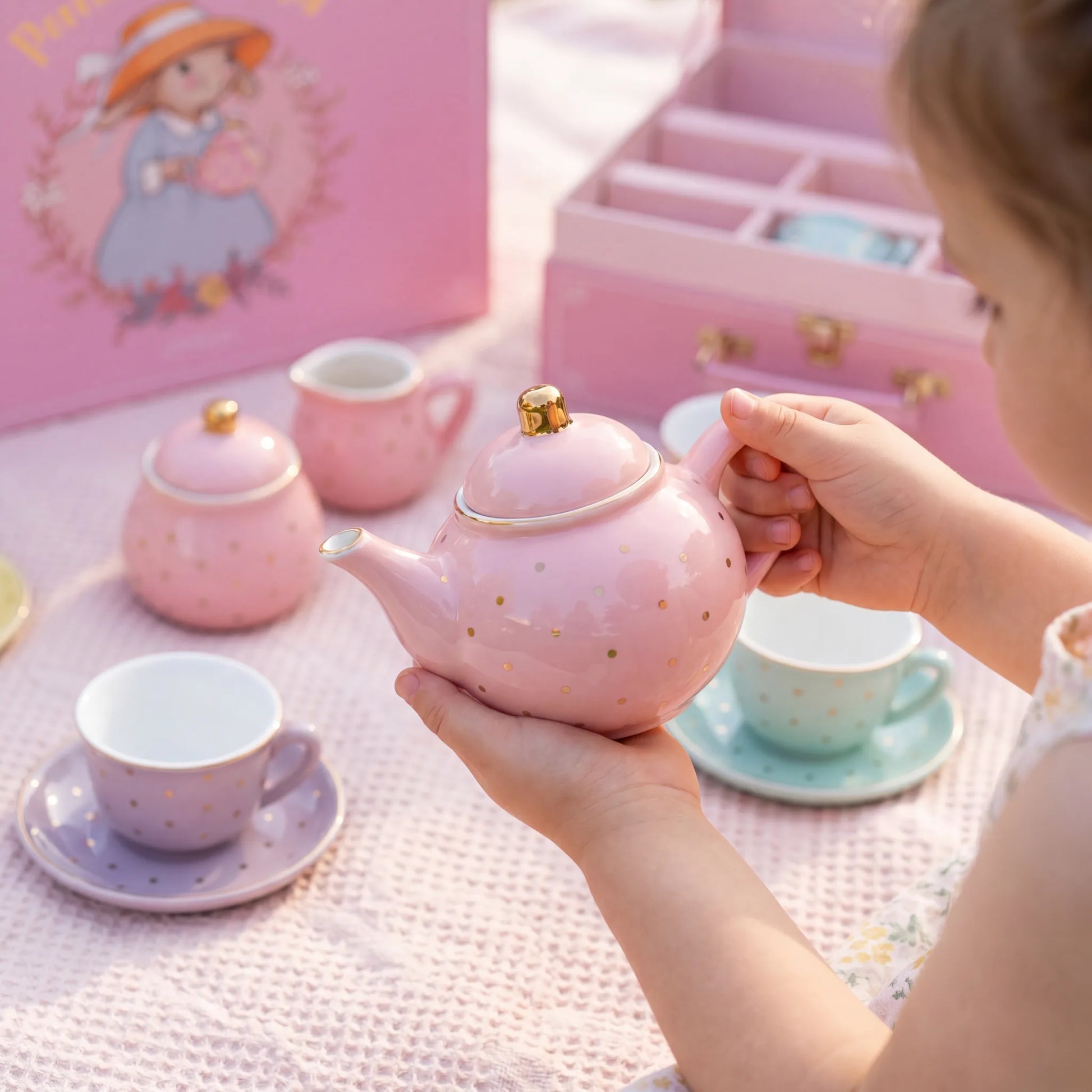 Child holding the pink polka dot porcelain teapot beside the pastel tea set and pink carry case